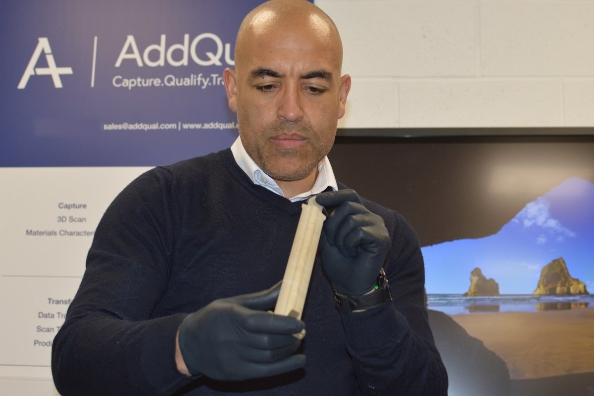 An AddQual engineer wearing protective gloves inspecting a long, pale component in front of an AddQual banner